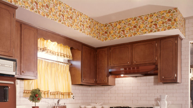 A vintage kitchen with brown cabinetry, a white tile backsplash, and a flower-patterned wallpaper border