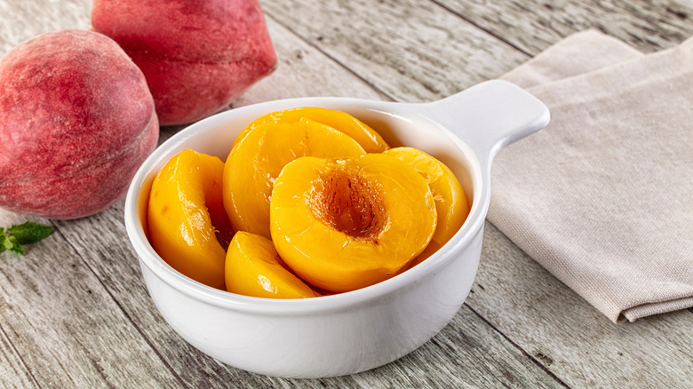Canned peaches sit in a white ramekin, near fresh peaches