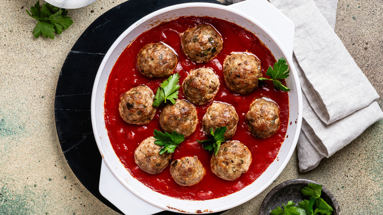 White bowl of meatballs in tomato sauce is topped with fresh flat leaf parsley