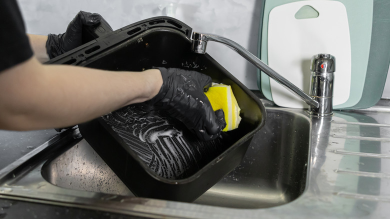 A person cleaning the air fryer tray in a sink with a yellow sponge