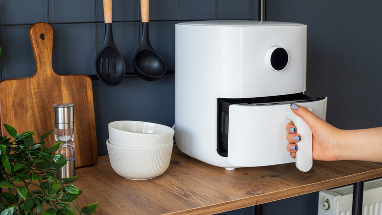 Hand pulling out the drawer of a white air fryer, on a kitchen counter with a cutting board and bowls nearby