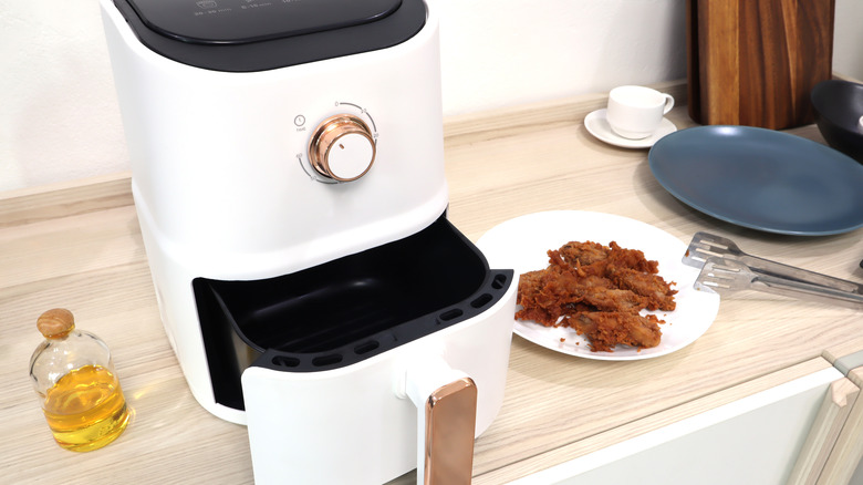 A white air fryer with a glass bottle of oil beside it and fried chicken